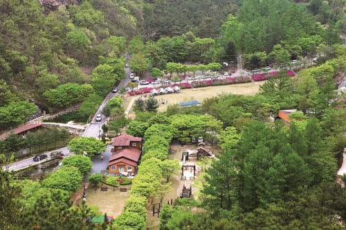 숲에 둘러싸인 고산자연휴양림의 잔디밭과 놀이터, 붉은 지붕 건물을 위에서 내려다본 모습