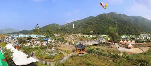 산자락 아래 개울을 따라 축제 천막과 부스가 늘어선 축제장 전경, 앞쪽에 정자와 개울을 건너는 다리
